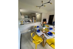 A modern kitchen with stainless steel appliances, a dining area with yellow chairs and blue plates, and a chandelier.