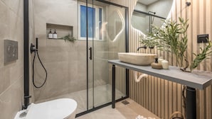A modern bathroom with a glass-enclosed shower, a toilet, a sink with a marble countertop, and a mirror with a unique curved design.