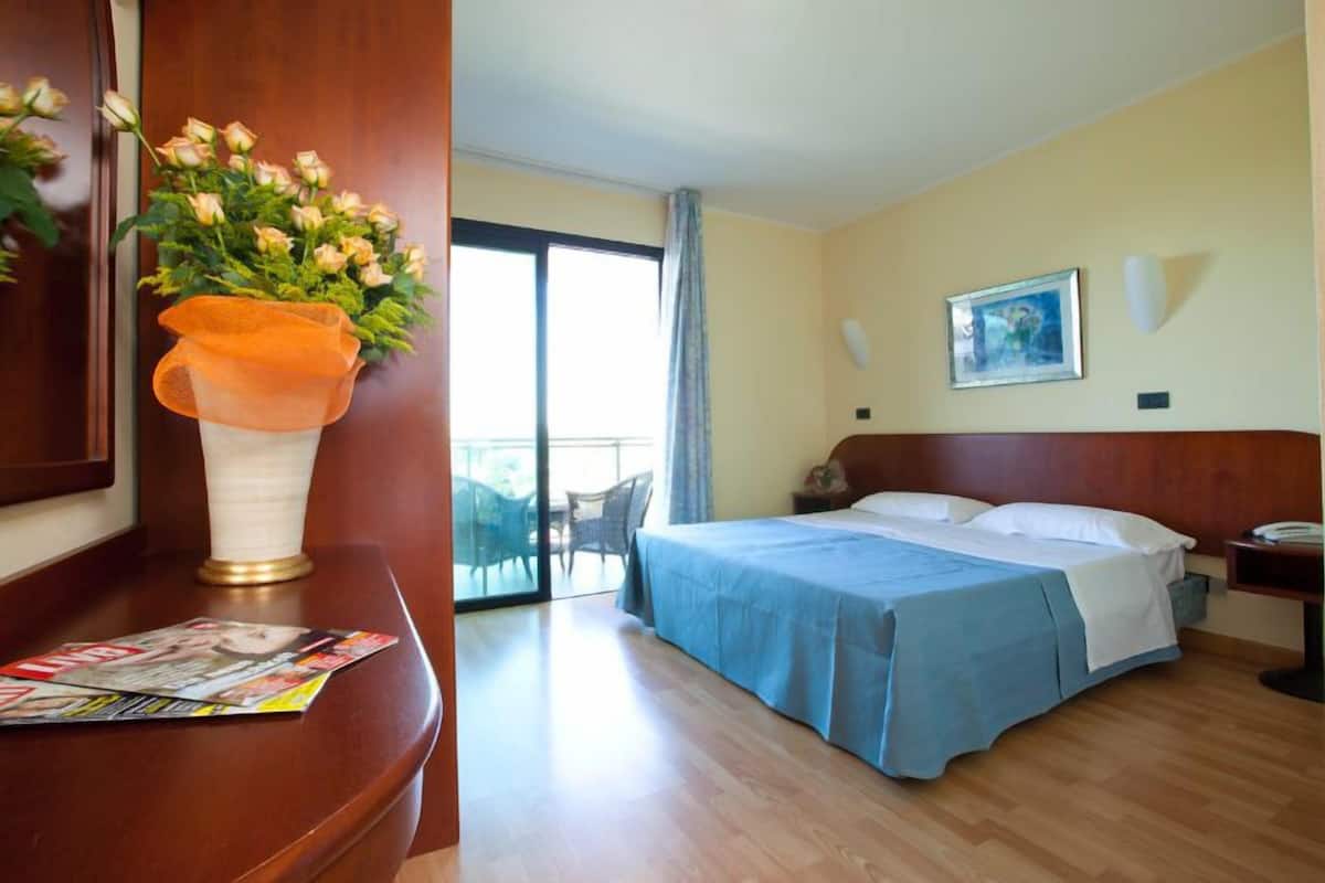 Une chambre d’hôtel avec un lit, une table de chevet, une chaise, une table avec un magazine, une fenêtre donnant sur un beau paysage et un vase rempli de fleurs.