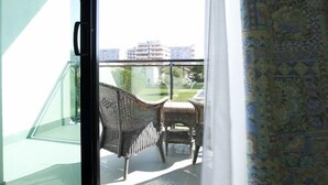 A room with a sliding glass door leading to a balcony with wicker furniture and a view of buildings and greenery. - Hotel Falcone (Lignano Sabbiadoro)