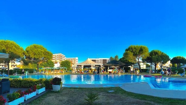 A swimming pool area with multiple pools, surrounded by greenery and buildings. - Hotel Falcone (Lignano Sabbiadoro)