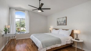 Una camera da letto con un letto, due comodini, una scrivania, una sedia, una ventola a soffitto e una finestra con tende.