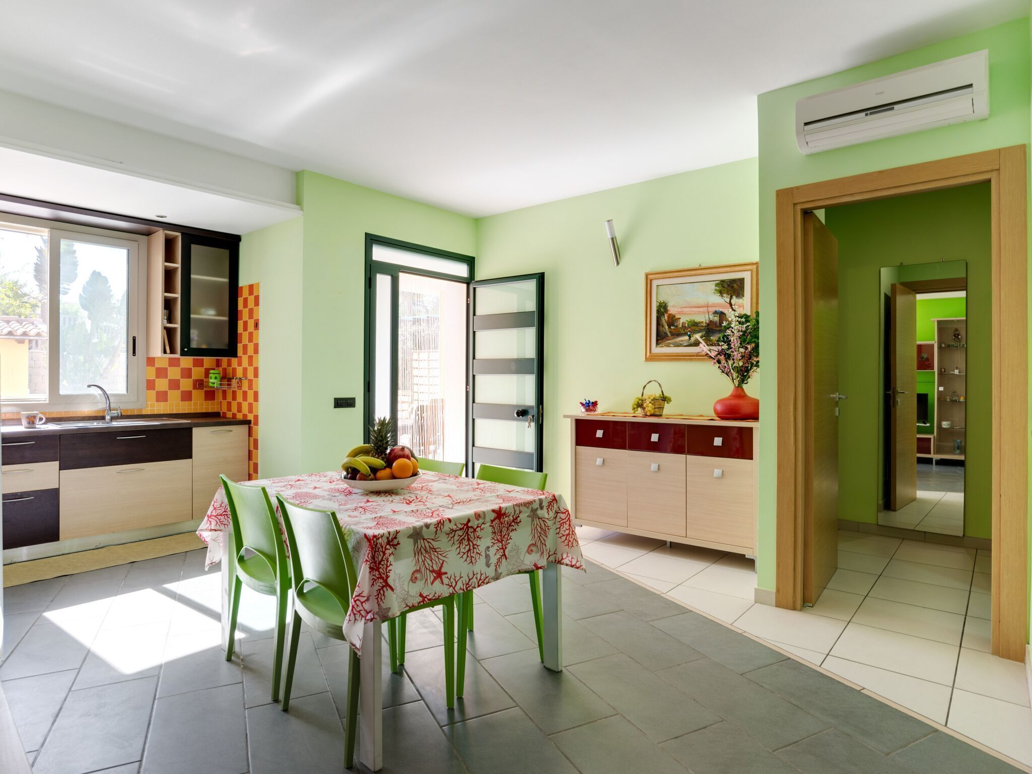 A bright kitchen with a dining table set for two, a fruit bowl, and a view of the outside.