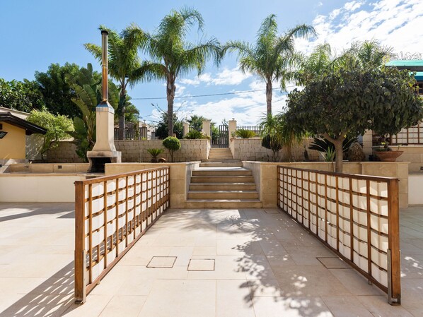 A paved pathway leading to a set of stairs, flanked by wooden fences and surrounded by palm trees and greenery.