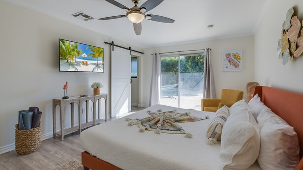 A bedroom with a bed, a ceiling fan, a TV, a window, and a door leading to a balcony.