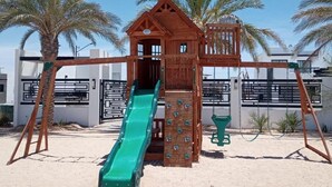 Eine Holzspielplatzkonstruktion mit Rutsche und Kletterwand, umgeben von Palmen und modernen Gebäuden.