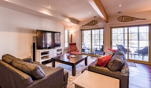 A modern living room with a large flat-screen TV, comfortable seating, and large windows offering a view of the outdoors.