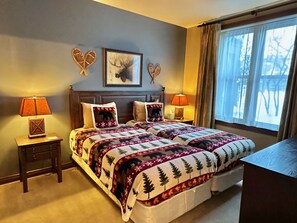 Un dormitorio con dos camas, cada una con una almohada temática, una mesita de noche con lámpara, un cuadro enmarcado en la pared y una ventana con cortinas.