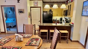 Un comedor acogedor con una mesa y sillas de madera, una cocina con armarios amarillos y un refrigerador, y una sala de estar visible a través de una puerta abierta.