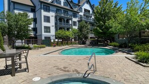 Un área de piscina con un jacuzzi, rodeada por un patio de ladrillo y árboles.