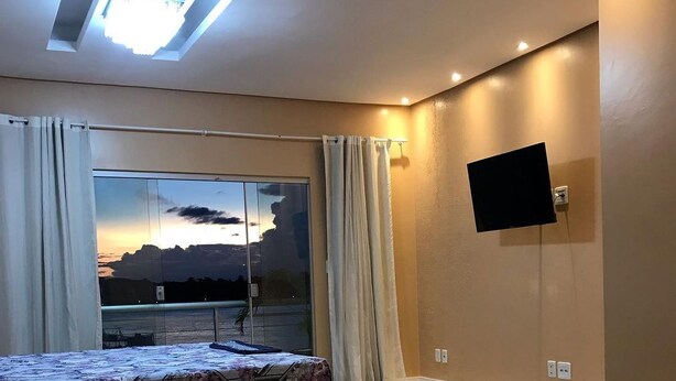 Un dormitorio con cama, televisor montado en la pared, un amplio ventanal con cortinas y vista al atardecer.