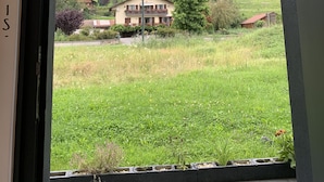 Vista a través de una ventana de una casa con balcón, rodeada de vegetación y un paisaje montañoso.