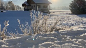 Una casa con techo nevado y un balcón, rodeada por un paisaje invernal y montañas al fondo.