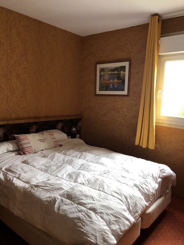 Un dormitorio con una cama grande, cabecera, un cuadro enmarcado en la pared y una ventana con cortinas.