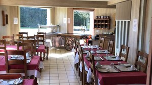 A restaurant with multiple tables and chairs, a kitchen area, and a view of trees outside. - Hotel Le Drakkar (Pont-Audemer)