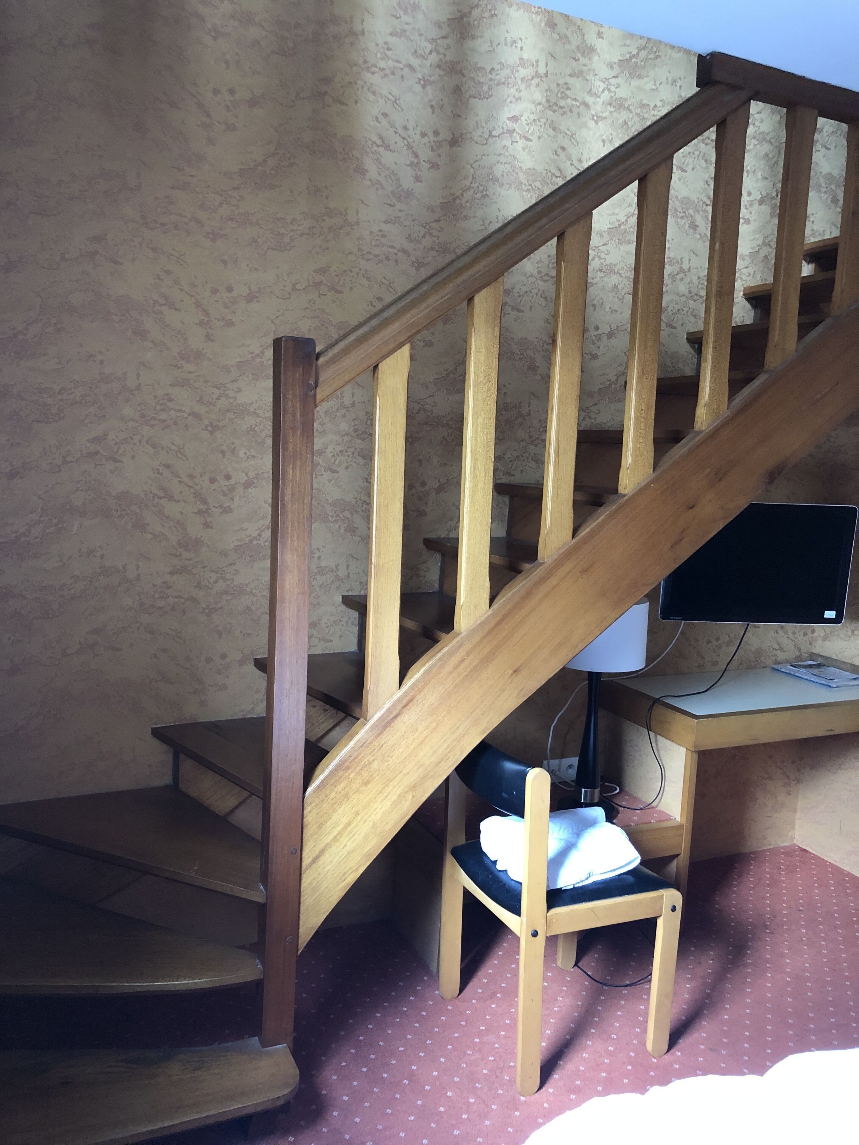 Una escalera de madera con un escritorio y una silla debajo, un monitor de computadora sobre el escritorio y una alfombra estampada en el suelo.