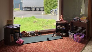 A room with a large window, a red carpet, a small table with books, and a toy shelf. - Hotel Le Drakkar (Pont-Audemer)