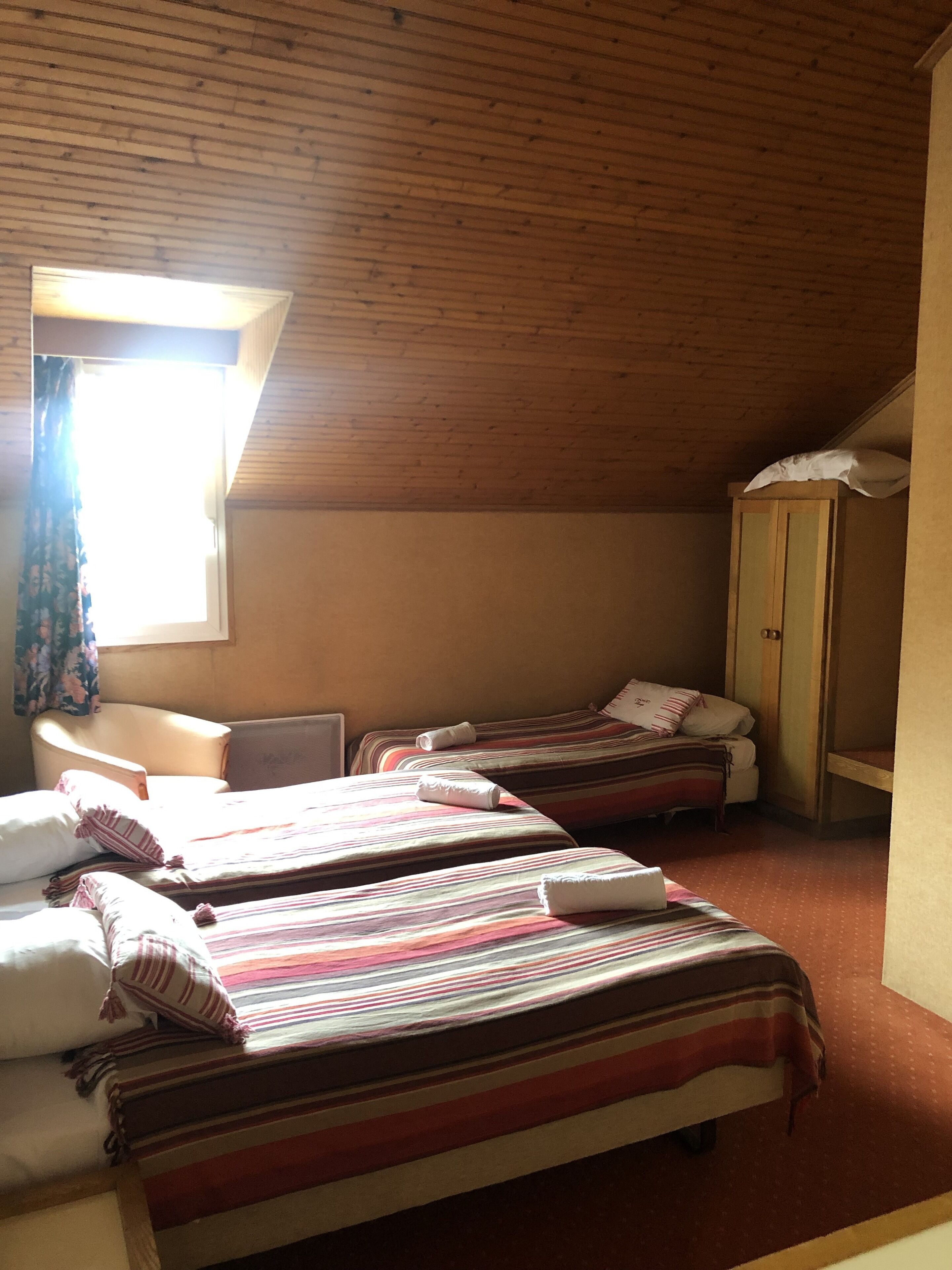 A room with two beds, a wooden ceiling, a window with curtains, and a wardrobe.