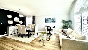 Een moderne woonkamer met een eethoek, een glazen salontafel en een groot raam met zonwering.