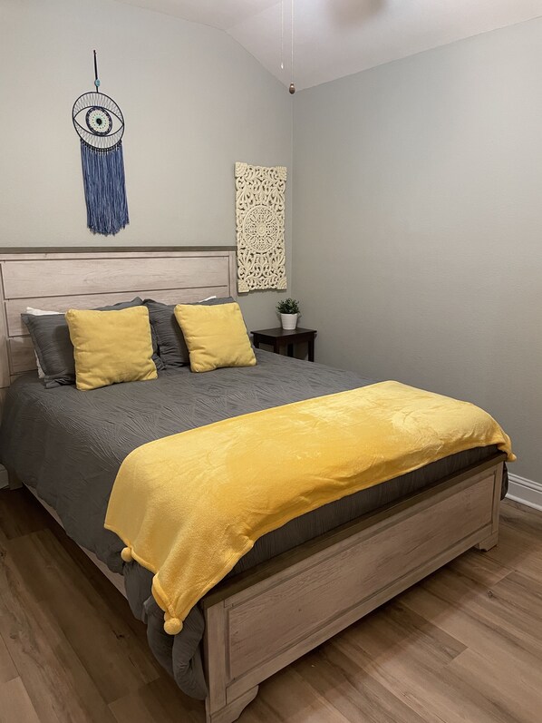 A bedroom with a wooden bed, a headboard, a wall hanging, and a small nightstand with a plant.