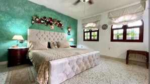 A bedroom with a tufted headboard, a bed with a white bedspread, two bedside tables with lamps, a decorative floral arrangement above the bed, and a window with a view of greenery. - 8MG-1 MANSION EN CARTAGENA CERCA AL MAR (Cartagena)