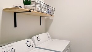 A laundry area with two white Amana washing machines, a wooden shelf with a potted plant and folded towels, and a metal basket. - New Condo Walkable to Downtown Bozeman (Bozeman)