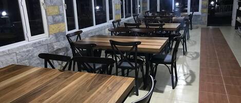 Un restaurante con varias mesas de madera y sillas negras, ventanas amplias a lo largo de una pared y piso de baldosas.