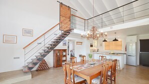Uma cozinha espaçosa com uma ilha central, uma área de jantar com mesa e cadeiras de madeira, e uma escada que leva a um andar superior.