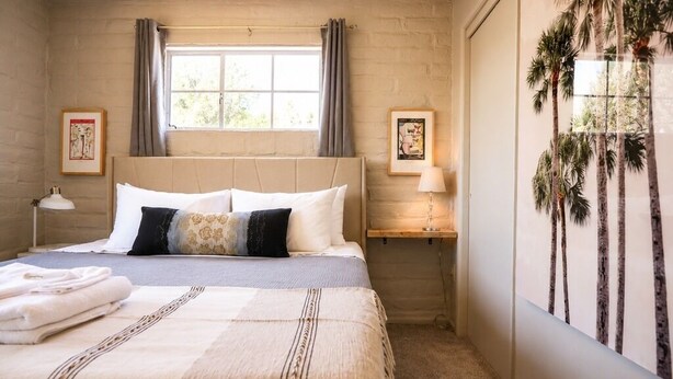Un dormitorio con cama, almohadas, una mesita de noche, una lámpara y un cuadro en la pared.