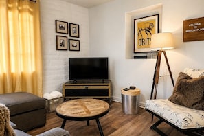 A living room with a flat-screen TV, a round wooden coffee table, a sofa, a lamp, and framed pictures on the wall. - Benton Place - A Stylish Southwest Retreat (Tucson)