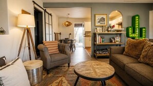 Una sala de estar con sofá, sillón, mesa de centro y lámpara. Se observa una estantería y una decoración con cactus.