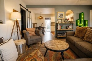 A living room with a sofa, armchair, coffee table, and lamp. There is a bookshelf and a cactus decoration.