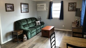 A living room with a green sofa, a wooden coffee table, a dining table with chairs, a ceiling fan, and two windows with curtains.