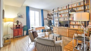 Un salon avec un plancher en bois, un canapé, un téléviseur, une bibliothèque et un coin repas comprenant une table et des chaises.