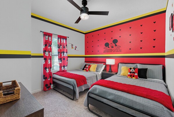 Quarto com duas camas, um ventilador de teto e uma parede vermelha decorada com o Mickey Mouse.