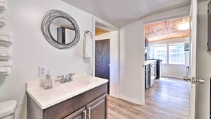 Un baño moderno con un tocador blanco, un espejo redondo y un piso de madera que conduce a una zona de cocina.