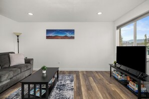 Un salon avec un canapé gris, une table basse, un meuble pour la télévision avec des jeux vidéo, et un tableau représentant un paysage.