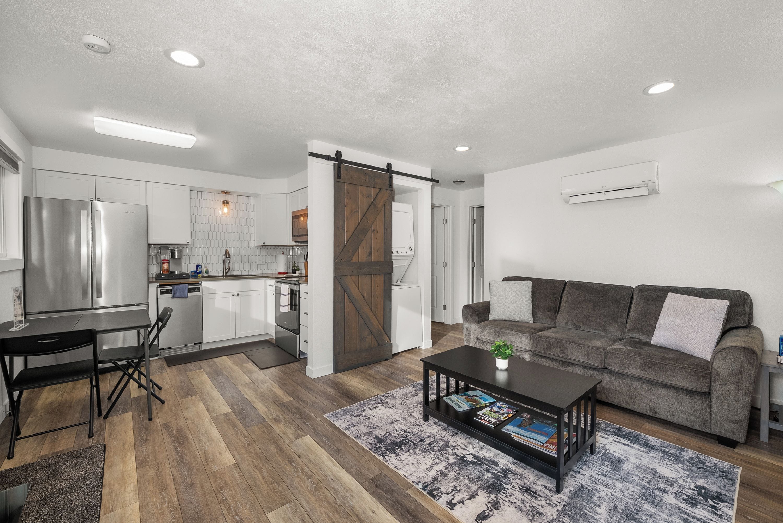 A modern kitchen with a stainless steel refrigerator, a dining area with a table and chairs, a gray sofa, and a coffee table with magazines.