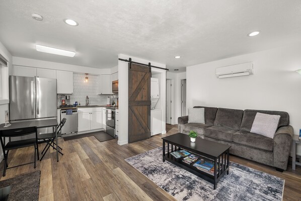 A modern kitchen with a stainless steel refrigerator, a dining area with a table and chairs, a gray sofa, and a coffee table with magazines.