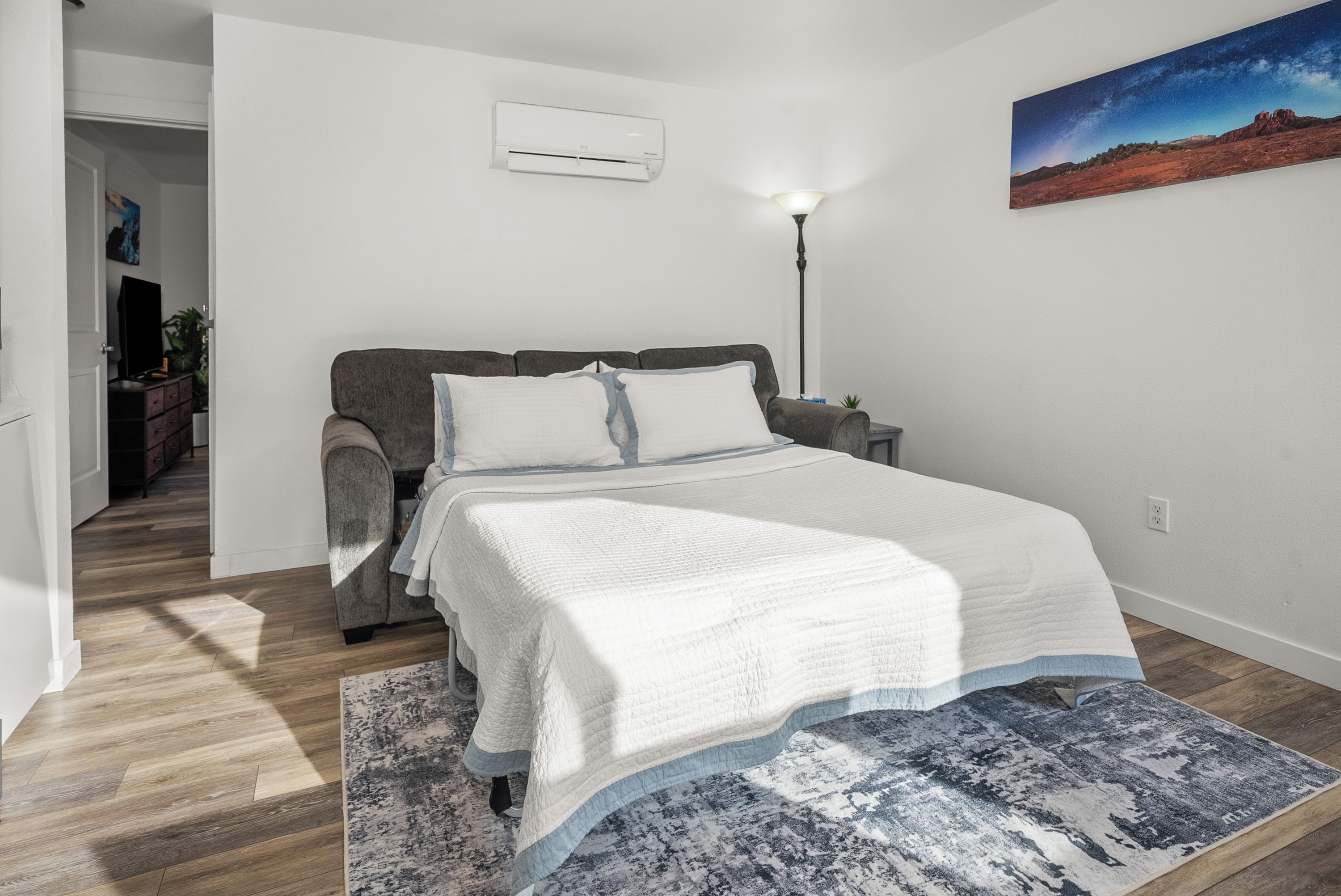 Ein Schlafzimmer mit einem Bett, einem Nachttisch, einer Lampe, einem Gemälde und einem Flur, der in einen anderen Raum führt.