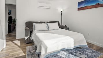 Un dormitorio con cama, mesita de noche, lámpara, un cuadro y un pasillo que conduce a otra habitación.