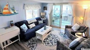 Una sala de estar con un sofá azul oscuro, un tapete estampado y una mesita auxiliar blanca. Hay una ventana con cortinas, una lámpara y vistas al balcón exterior.