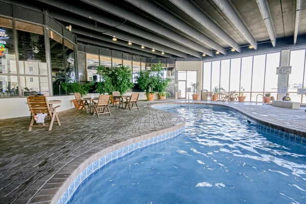 An indoor swimming pool with tiled edges, surrounded by wooden chairs and potted plants.
