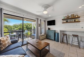 A modern living room with a sofa, coffee table, and bar stools, a TV mounted on the wall, and a balcony with a view of greenery. - Oceanfront Condo - Rare King Bed + Bikes & Walk to Coligny (Hilton Head Island)