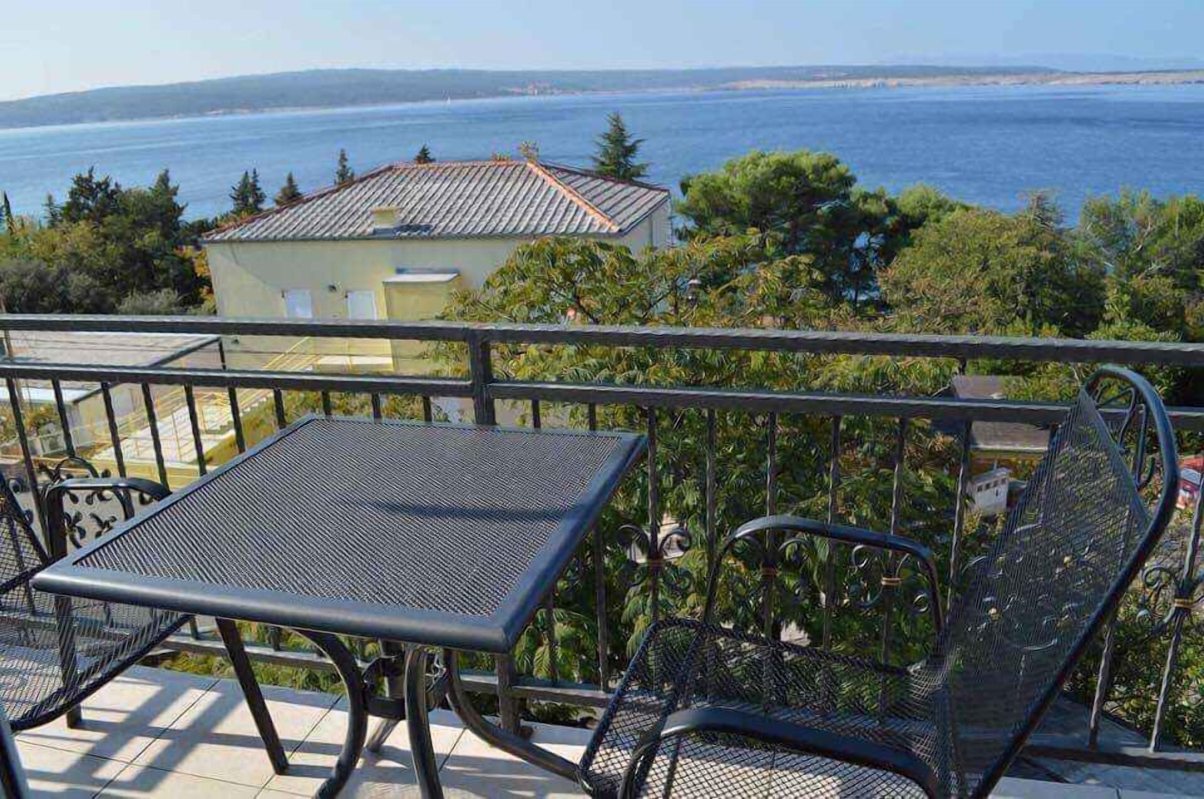 A balcony with a metal table and chairs, overlooking a body of water and a building with a tiled roof.