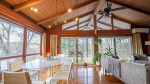 Una habitación amplia con techo de madera, ventanas grandes, una mesa de comedor con sillas, un sofá y un ventilador de techo.