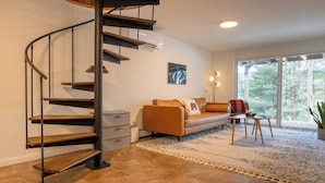 Una sala de estar moderna con una escalera espiral, un sofá de cuero, una mesita y un ventanal con vistas a árboles.