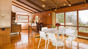 Una cocina amplia con vigas de madera, un comedor con sillas blancas y ventanas grandes que ofrecen vistas al exterior.
