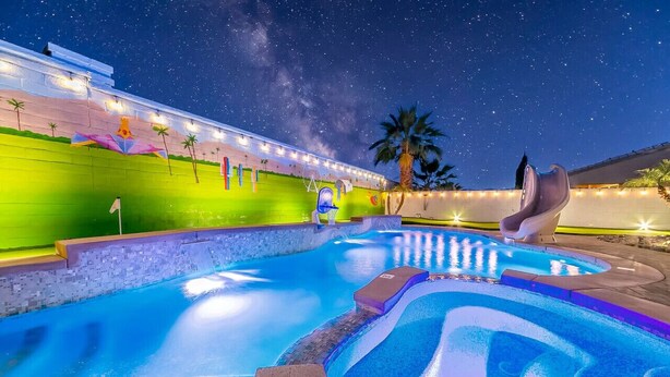 Ein Nacht-Schwimmbad mit Wasserrutsche, Palmen und einem Gebäude mit beleuchteten Fenstern.