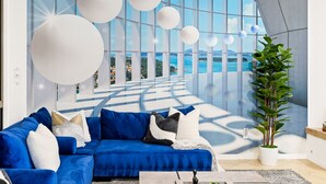 Ein modernes Wohnzimmer mit einem blauen Sofa, weißen, kugelförmigen Lampen und einem großen Fenster mit Meerblick.